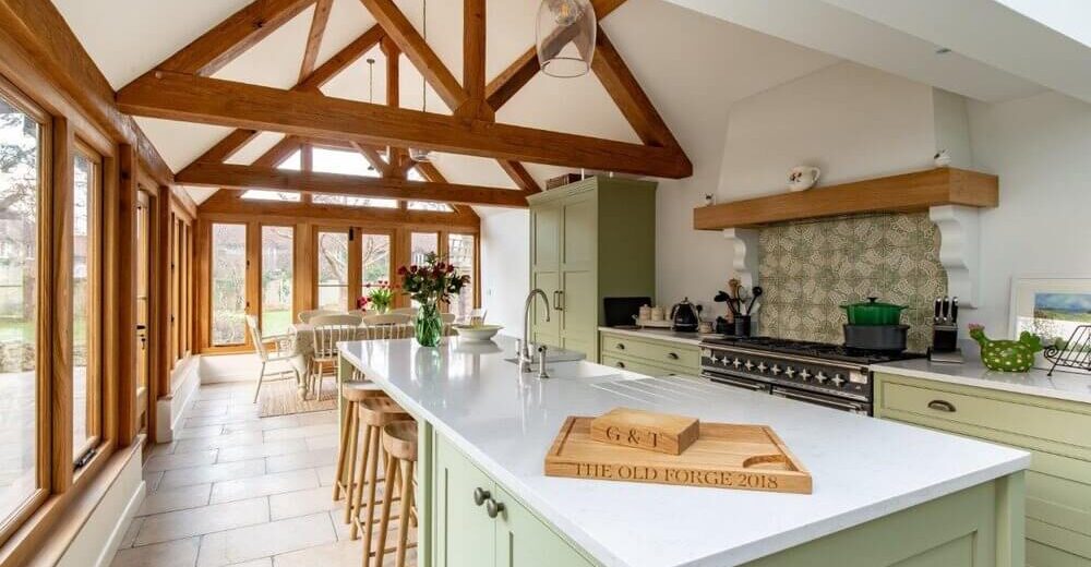 A spacious kitchen with a large island featuring mint green cabinets and wooden bar stools. The ceiling has exposed wooden beams, and large windows provide natural light. A rustic cutting board and a patterned backsplash add charm.