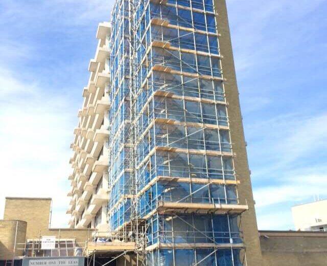 A tall building covered in scaffolding with blue netting, set against a clear sky, signals the progress of Kent Builders. Several vehicles are parked nearby, including a black car and a white truck, as a few people bustle around the base of the structure.