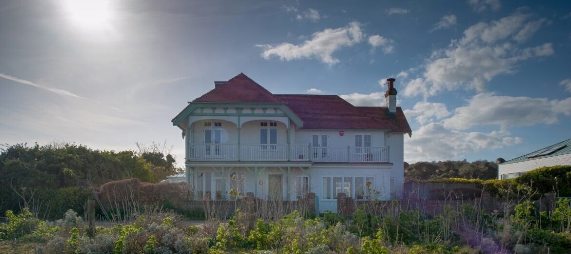 A charming two-story house with a red roof stands amidst overgrown greenery under a partly cloudy sky. The sun shines brightly, casting a warm light on the scene. The landscape is rustic and serene.