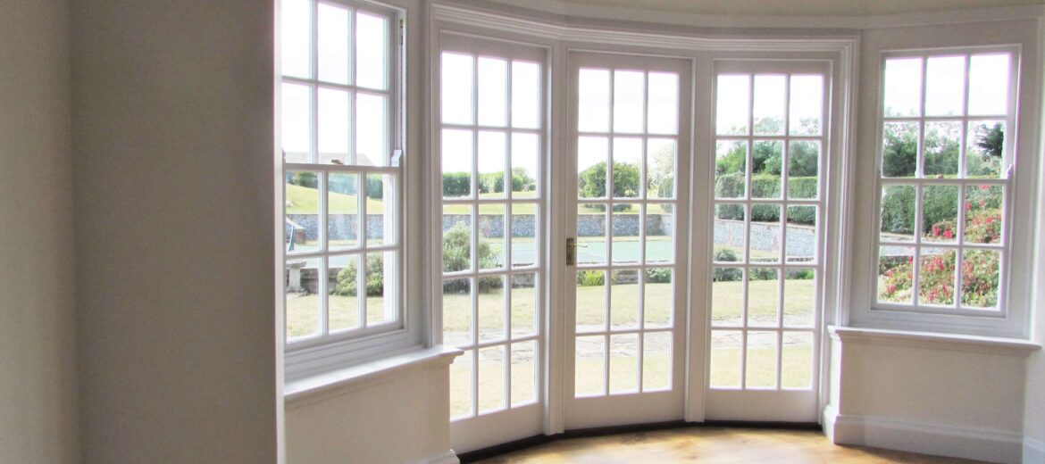 A bright, empty room with wooden flooring features a large bay window with multiple glass panes. The window offers a view of a garden with grass, shrubs, and trees under a clear sky.