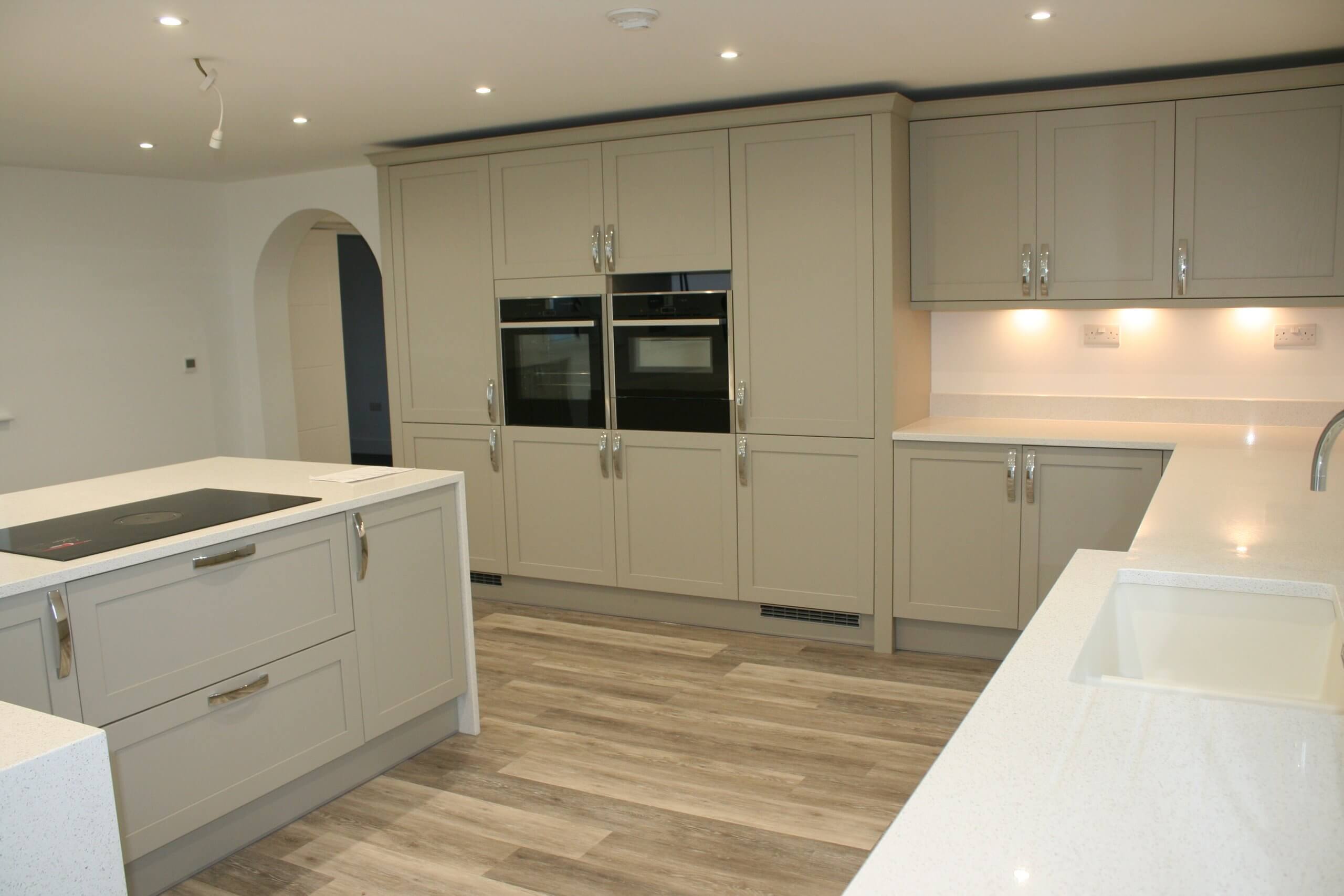 Modern kitchen with light wood flooring and cream cabinets. Features a large island with an electric stovetop, built-in ovens, and sleek countertops. Soft under-cabinet lighting enhances the clean, minimalist look.