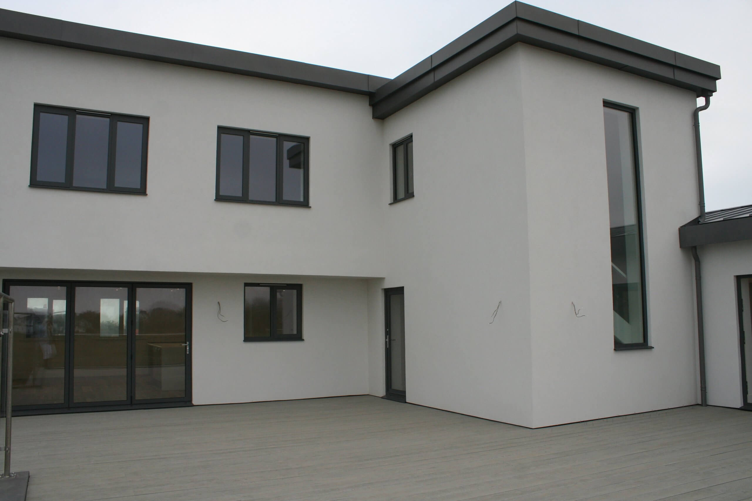 A modern two-story white house with dark-framed windows and a minimalist design. The building features a flat roof and large glass doors opening onto a wooden deck. The setting appears to be overcast.
