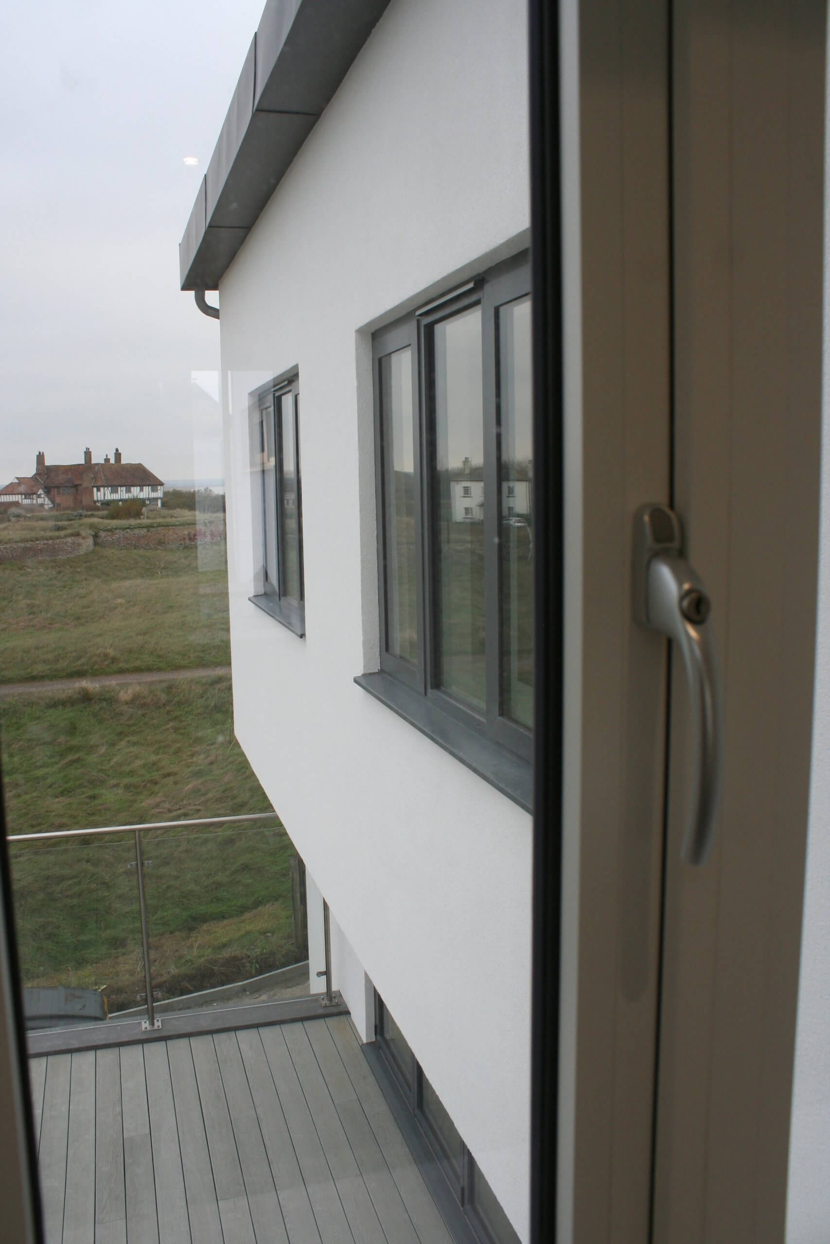 View from a window showing a modern white building with gray-framed windows and a glass balcony. In the background, grassy fields and a few houses can be seen under a cloudy sky.