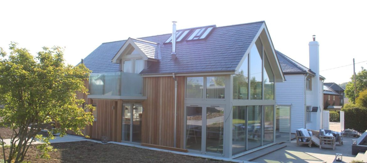 Modern house with a mix of glass and wooden panels, featuring a large slanted roof and multiple skylights. The exterior includes a spacious patio and surrounding greenery under a clear sky.