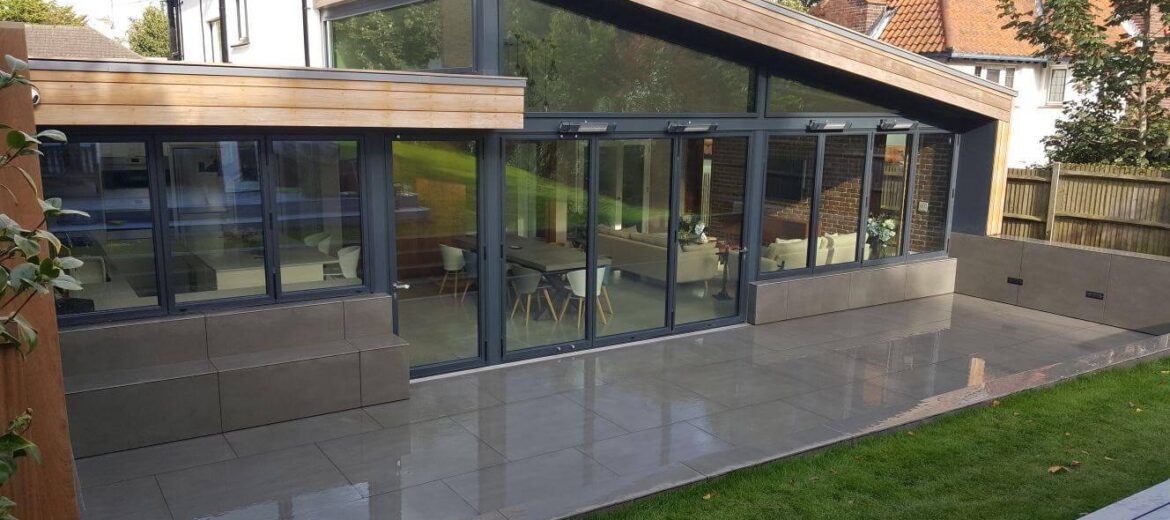 Modern house with a glass-fronted extension featuring a sloping roof, large windows, and sliding doors. The patio is tiled, and a neatly manicured lawn is in the foreground. The exterior walls are beige and gray.
