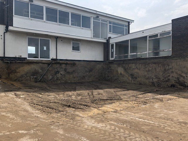 A large excavation site in front of a two-story building with white siding and large windows. The ground is cleared and partially dug out, surrounded by exposed soil, with an unfinished appearance under an overcast sky.