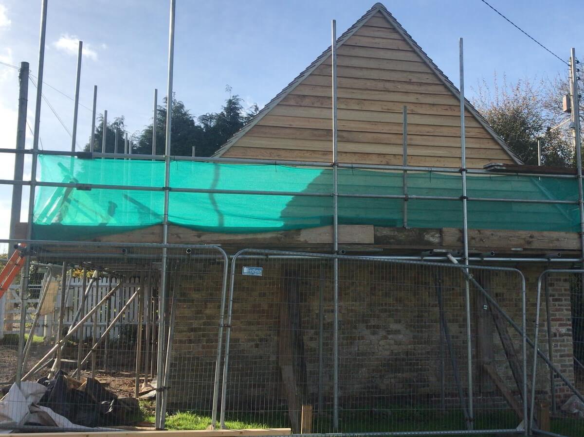 A building under construction with wooden siding and a brick base, surrounded by metal scaffolding. A green tarp covers part of the structure, with a clear blue sky in the background. Piles of material are visible on the ground.
