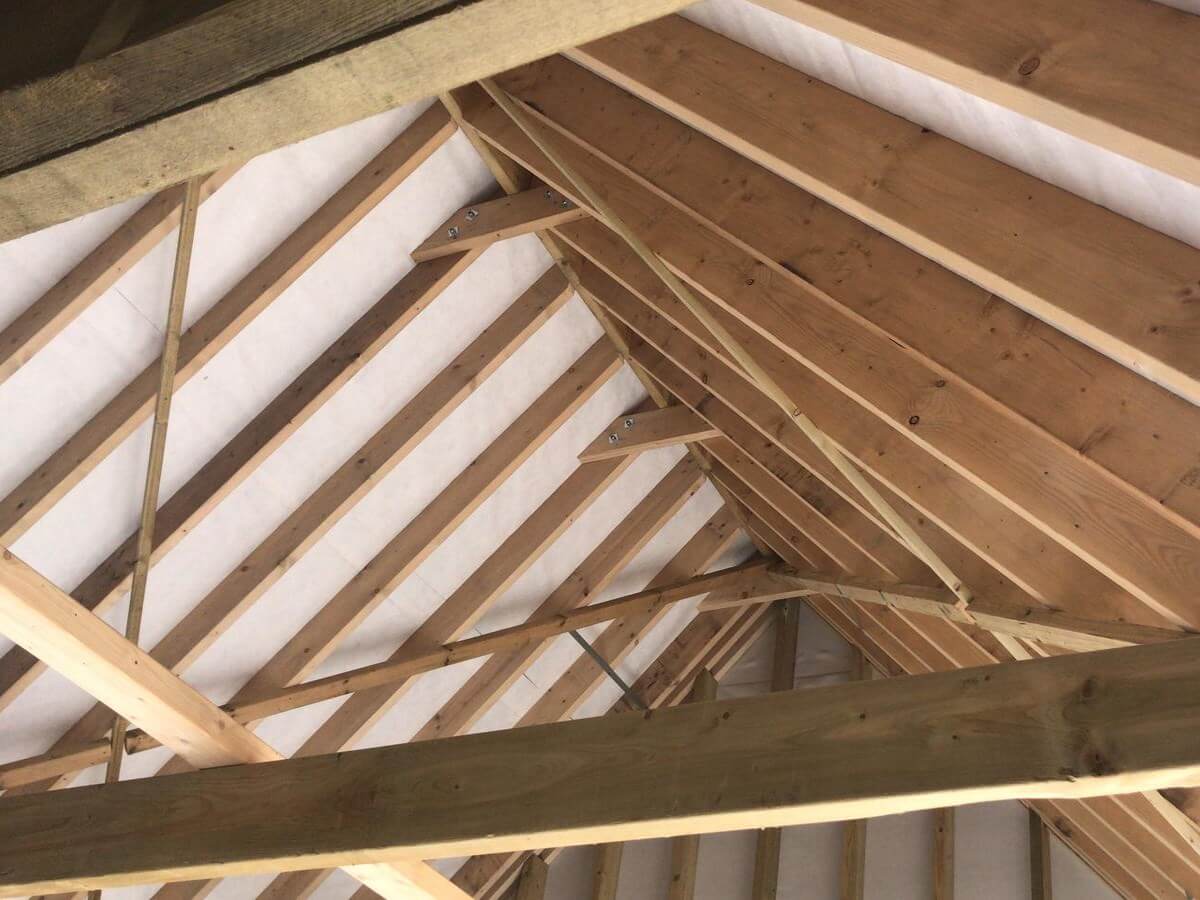 Wooden roof trusses are shown from inside, displaying beams and rafters in a triangular pattern. The wood is light-colored and forms a symmetrical structure, supporting the roof above, with some daylight visible between the beams.
