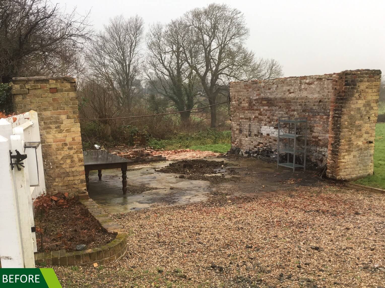 A weathered brick wall and a pile of rubble stand on a gravel surface, adjacent to a grassy field. Bare trees are visible in the background with an overcast sky. A BEFORE label is in the bottom left corner.