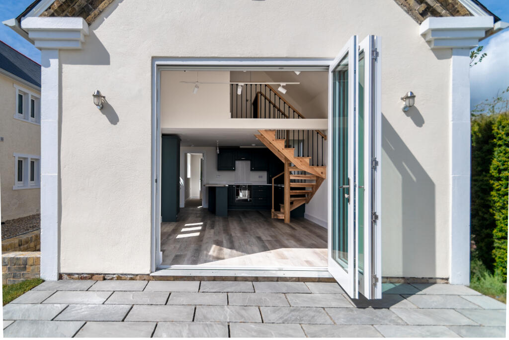 A modern house with open glass doors shows a spacious interior, wooden staircase, dark kitchen cabinets, and lots of natural light. A stone patio is in the foreground.