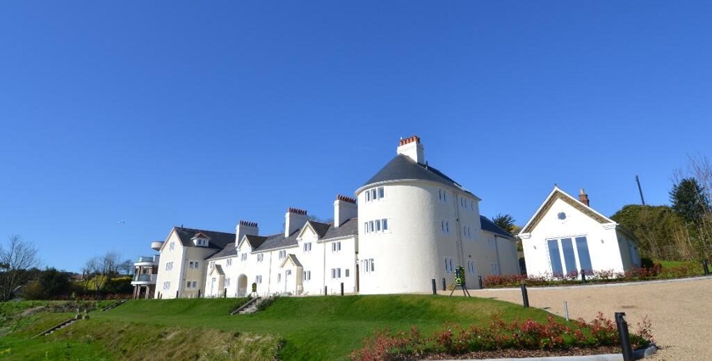Large white building with a round central tower, chimneys, and multiple pitched roofs, set on green lawns under a clear blue sky, with a gravel driveway in the foreground.
