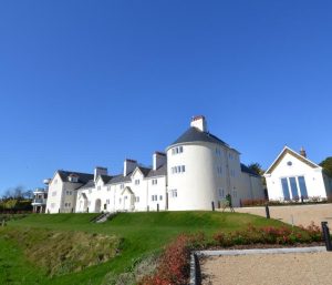 Large white building with a round central tower, chimneys, and multiple pitched roofs, set on green lawns under a clear blue sky, with a gravel driveway in the foreground.