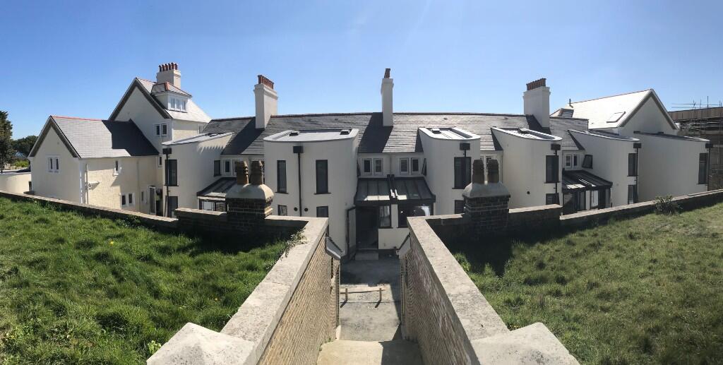 A row of modern, white houses with multiple chimneys, set behind a stone walkway and green grassy area, under a clear blue sky.