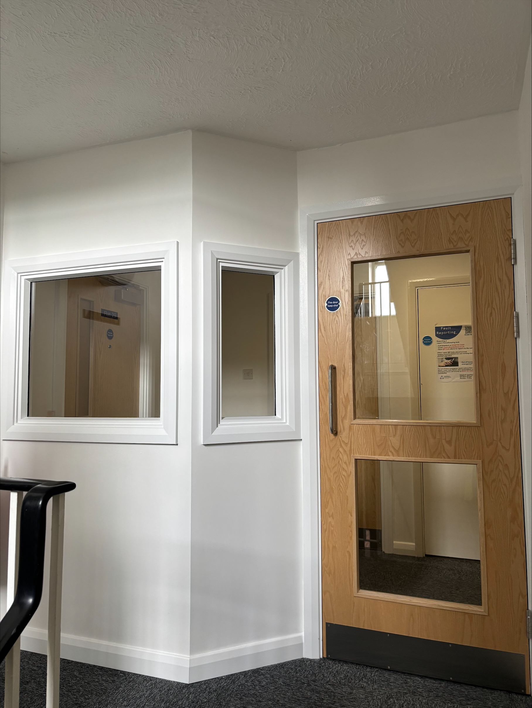 A white hallway with two interior windows and a wooden door with three glass panels. The door and windows have white trim, and there is a staircase railing visible on the left side.