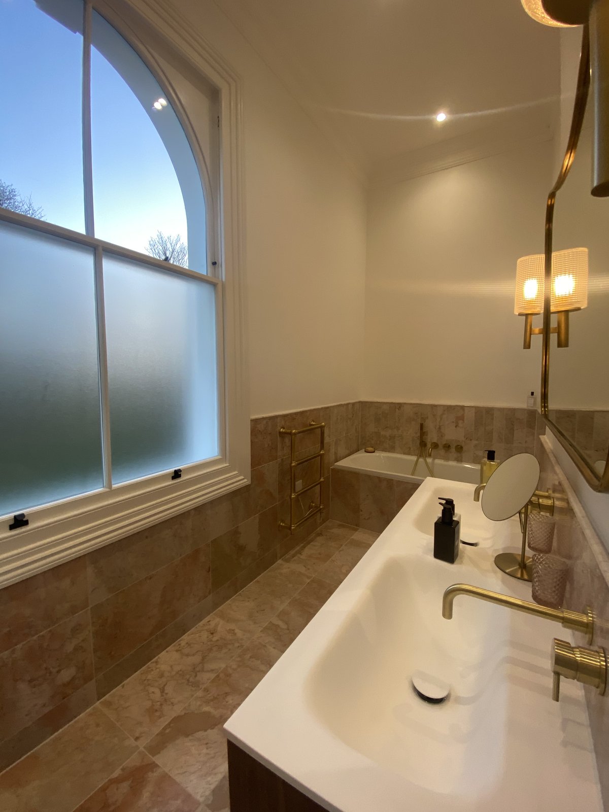 A modern bathroom with a large frosted window, a double sink with gold fixtures, a wall mirror, a towel rack, and a bathtub with beige marble tile. Warm lighting creates a cozy atmosphere.