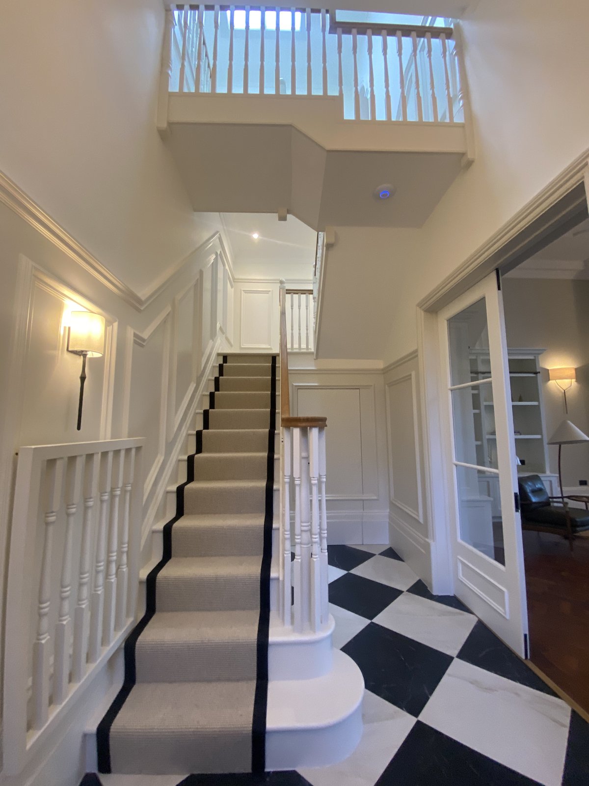 A modern staircase with a beige carpet and black runner leads to an upper floor. The hallway features white walls, checkered black-and-white flooring, glass doors, and white railings.