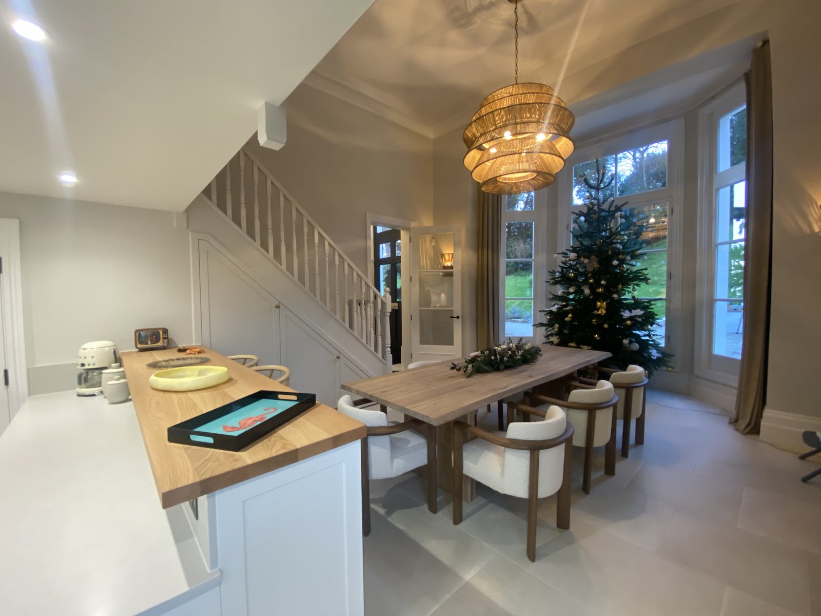 Modern dining room with a wooden table, eight chairs, Christmas tree, and hanging light fixture. Stairs and large windows are visible; kitchen counter is in the foreground with trays and appliances.