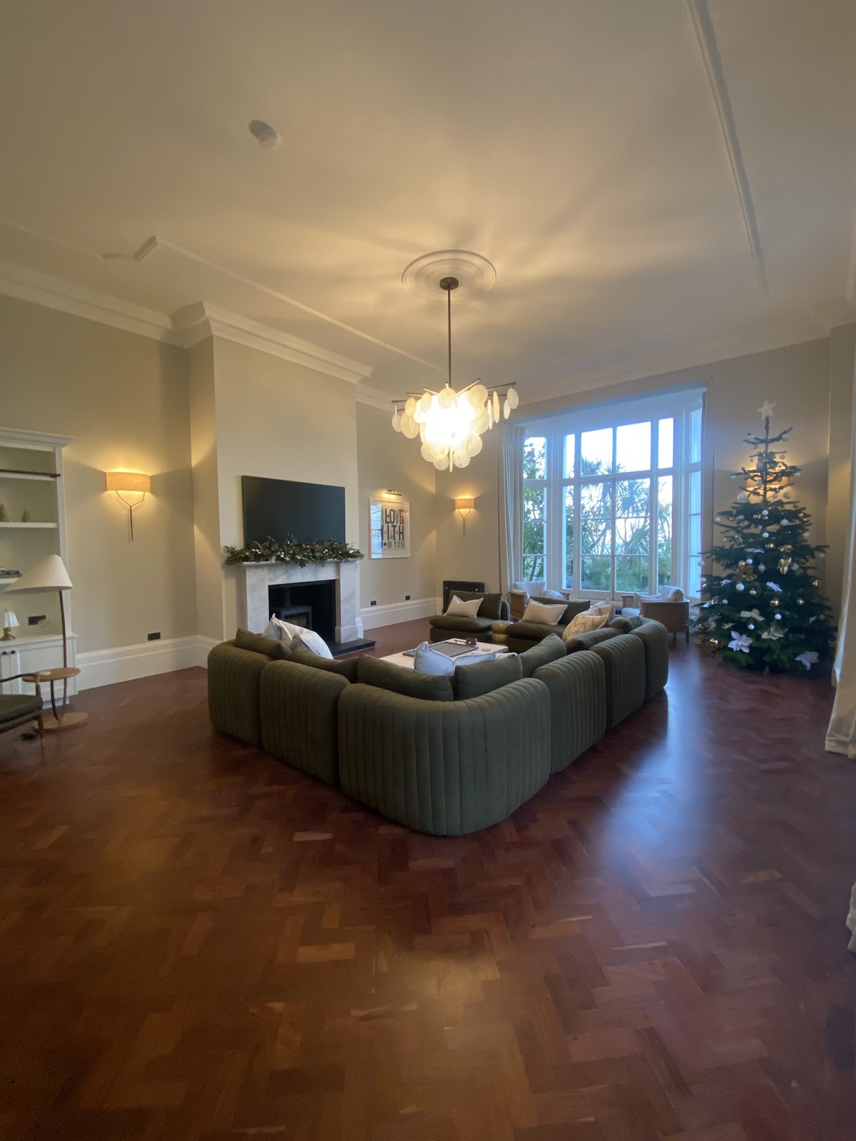 Spacious living room with a green sectional sofa, modern chandelier, fireplace, decorated Christmas tree, and large windows letting in natural light.