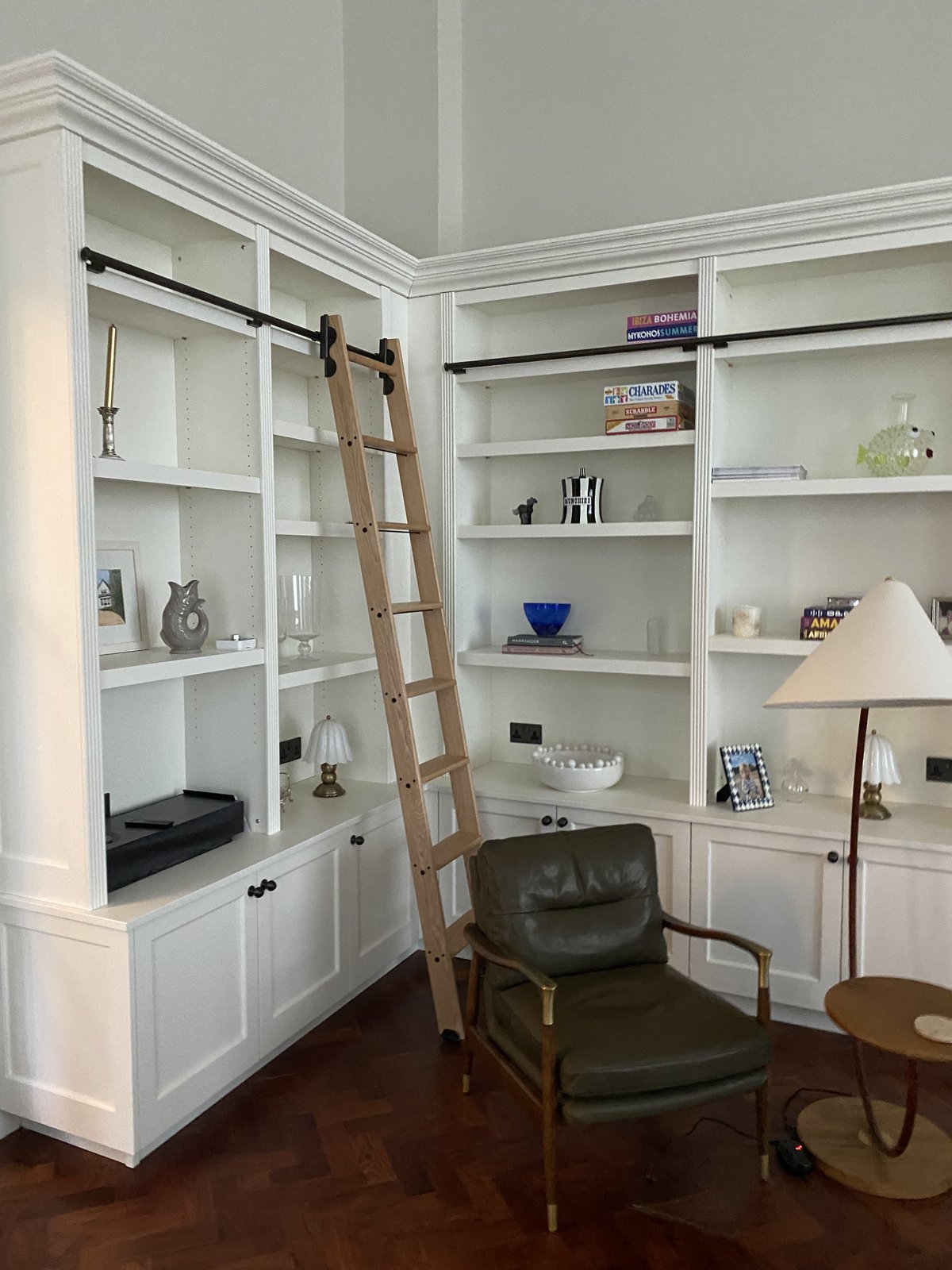 A wooden ladder leans against white bookshelves with various decor items, books, and a turntable. An olive green armchair and a small lamp-topped table sit nearby on a herringbone wood floor.