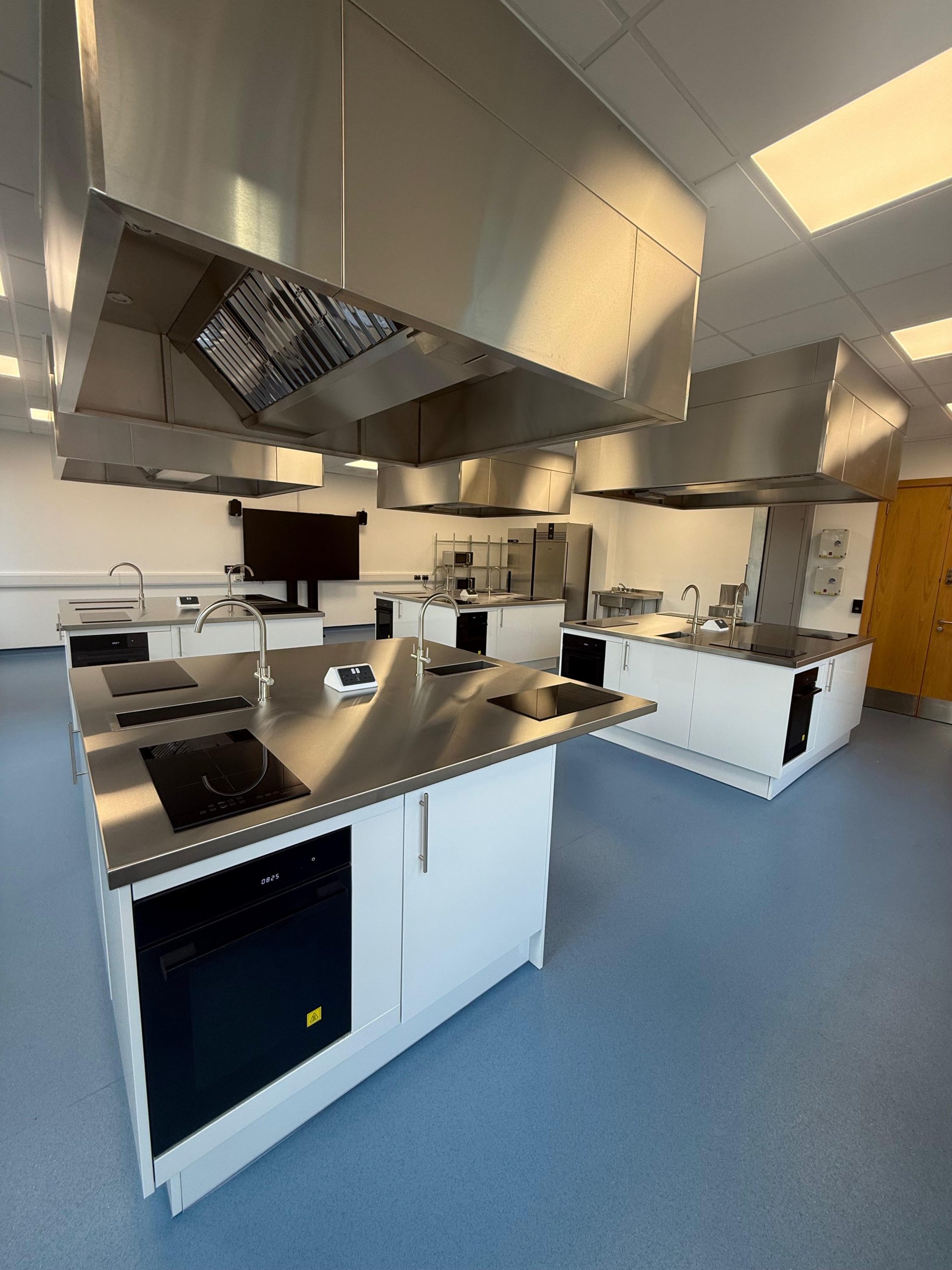 A modern, spacious kitchen classroom with stainless steel workstations, stovetops, ovens, sinks, and large ventilation hoods, under bright ceiling lights and a blue floor.