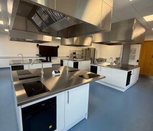 A modern, clean commercial kitchen with stainless steel workstations, induction cooktops, ovens, sinks, and overhead ventilation hoods on a blue floor. The room is well-lit with large windows and white walls.