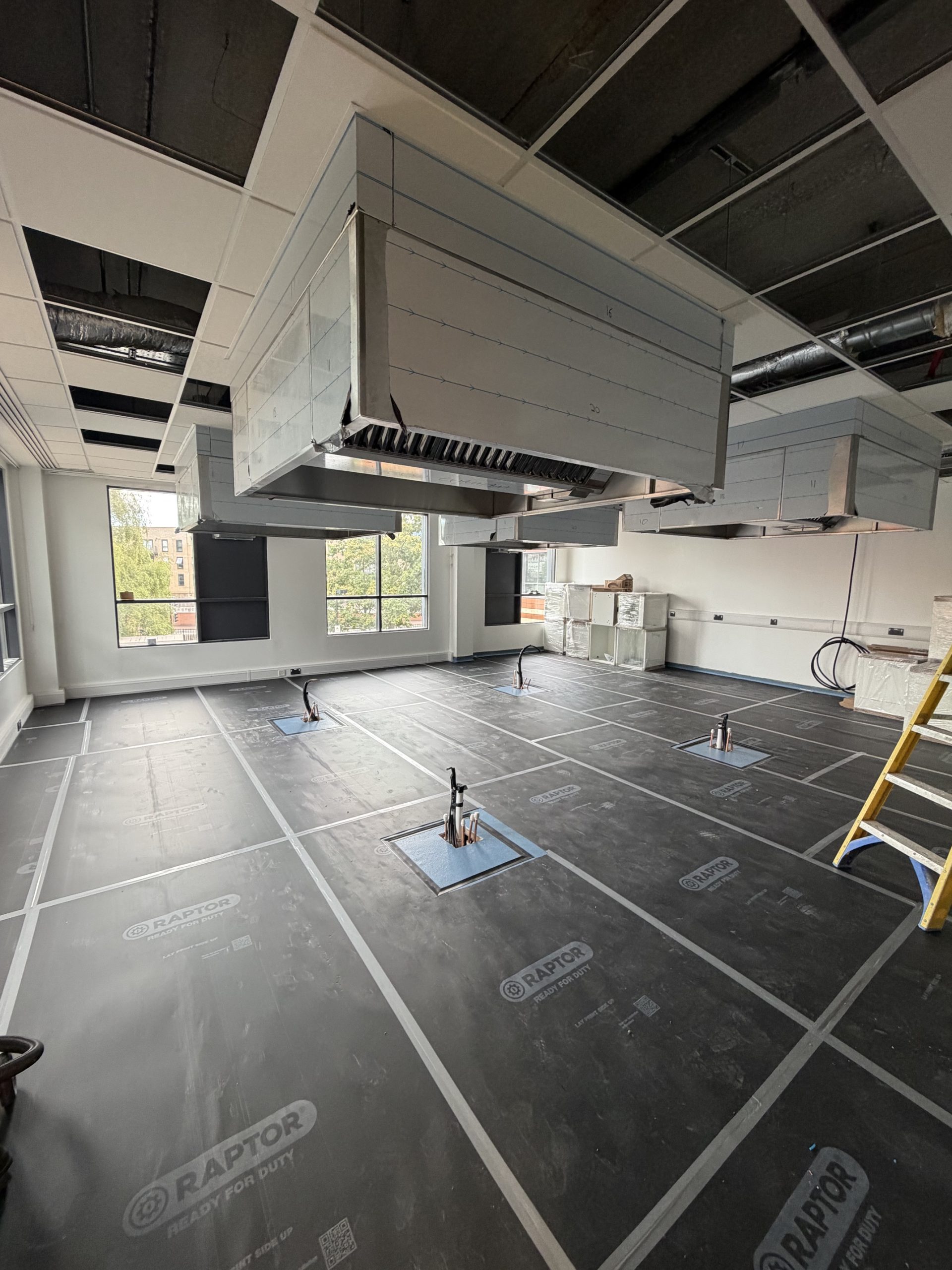 A partially constructed commercial kitchen with exposed vents, unfinished ceiling, covered floor, and pipes protruding where appliances will be. Large windows let in natural light. A yellow ladder stands to the right.
