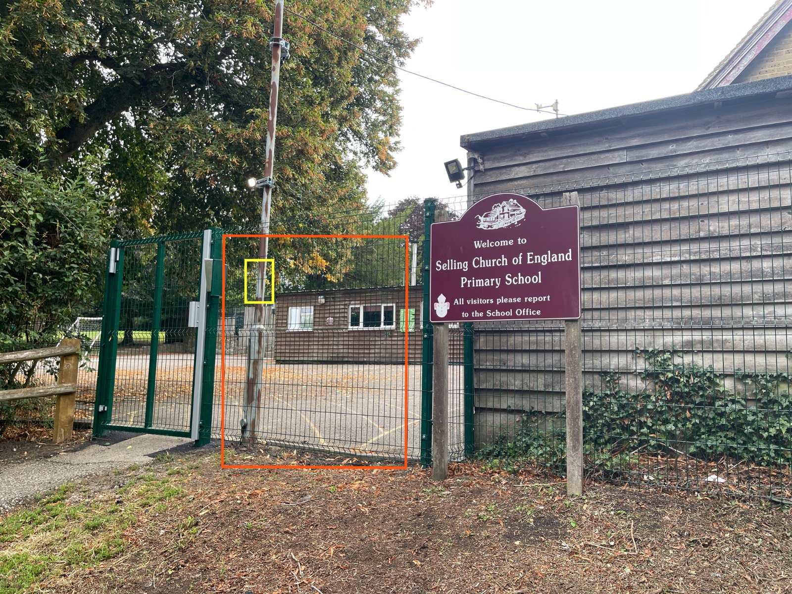 Entrance gate to a school with a purple sign reading, Selling Church of England Primary School. The gate is green metal, partially open, next to a wooden building and trees in the background.