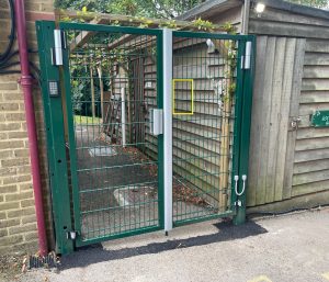 Green metal gate with keypad and lock, attached to a wooden building. Theres a sign on the building reading The Volunteers Society. Trees and a pathway are visible behind the gate.
