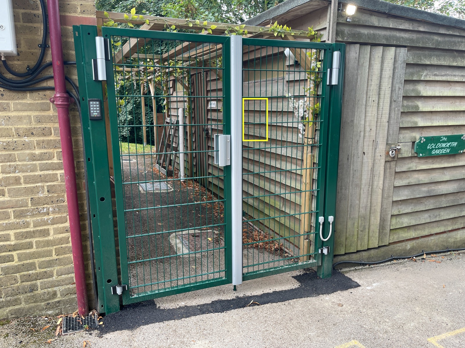 Green metal gate with keypad and lock, attached to a wooden building. Theres a sign on the building reading The Volunteers Society. Trees and a pathway are visible behind the gate.