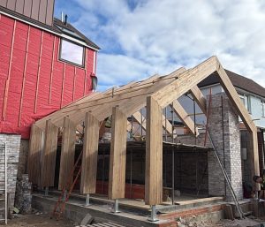 A partially constructed wooden house frame next to a red building, with scattered bricks, a ladder, and a traffic cone in the foreground under a cloudy sky.