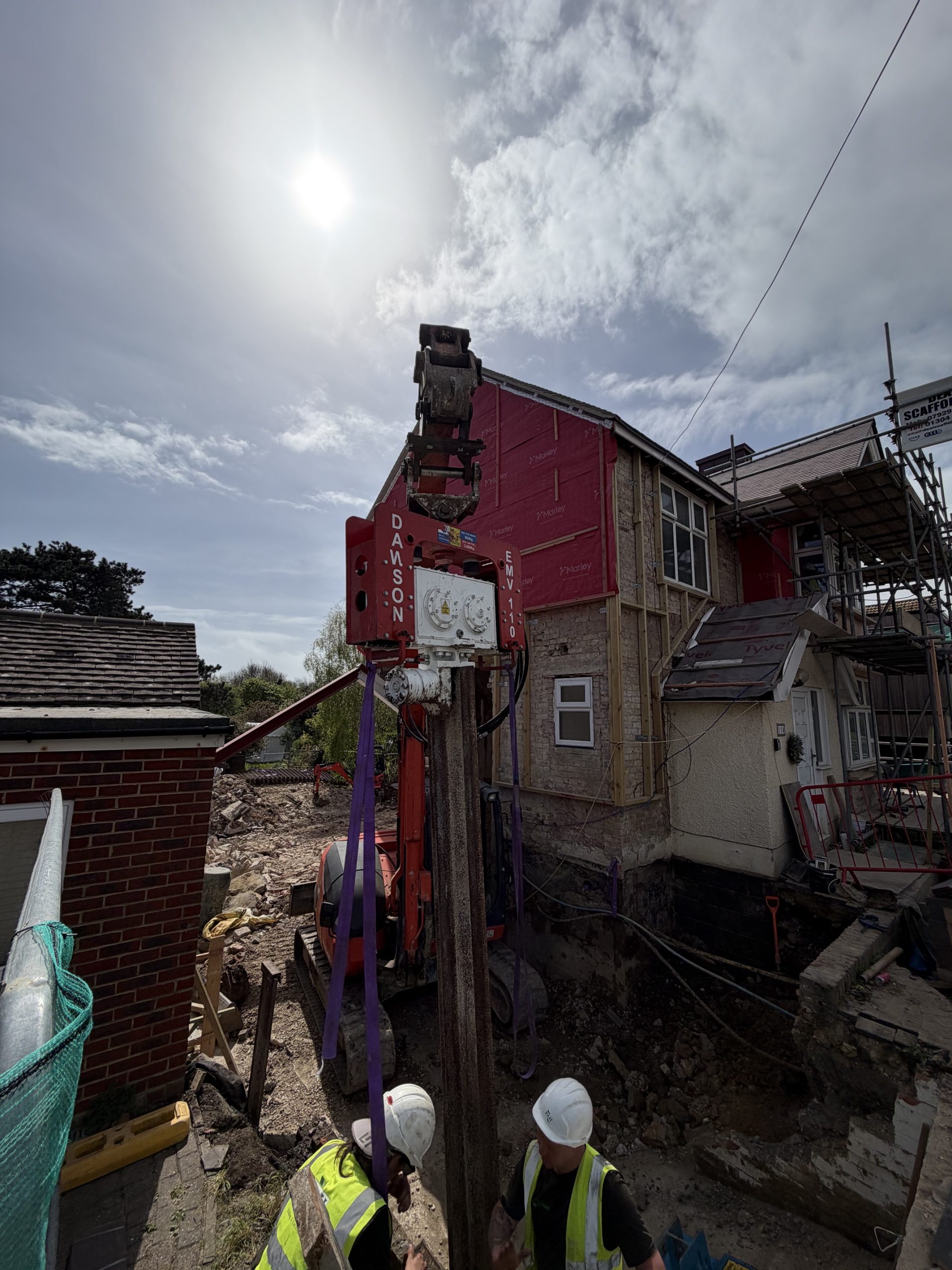 A construction site with workers in helmets operating machinery near a partially demolished house under a bright sun and cloudy sky.