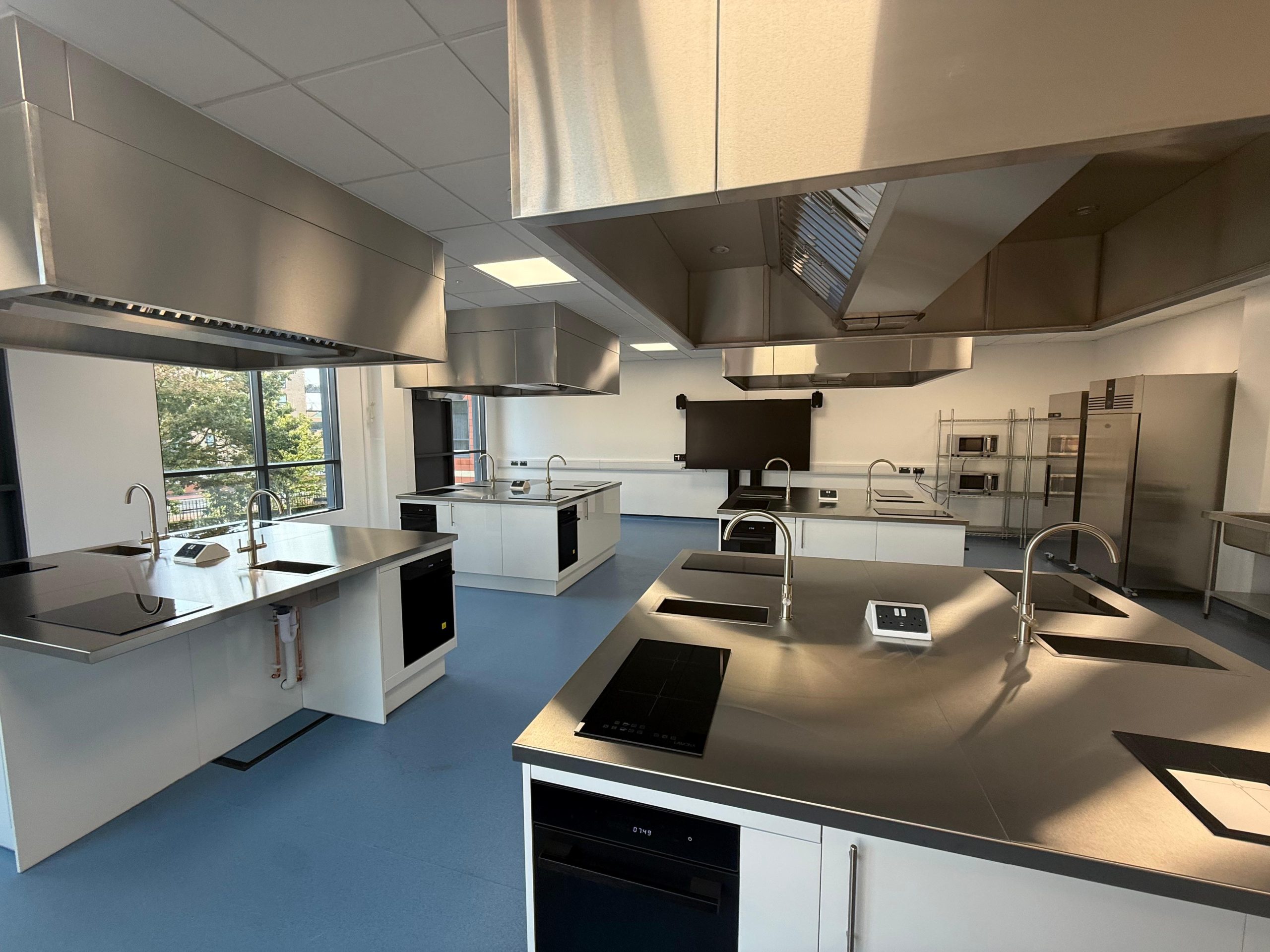 A modern, spacious kitchen classroom with multiple cooking stations, stainless steel appliances, induction cooktops, sinks, and large windows letting in natural light.