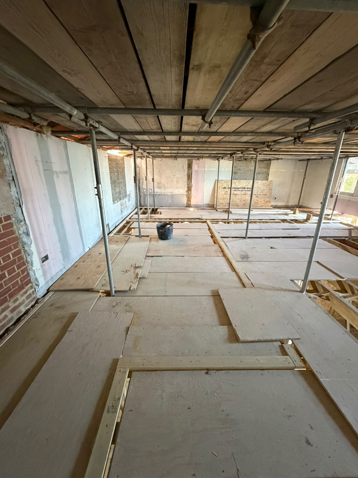 View of a building under renovation, with exposed wooden scaffolding and floorboards, unfinished walls, and a black bucket placed near the center of the room. Natural light enters through windows in the background.