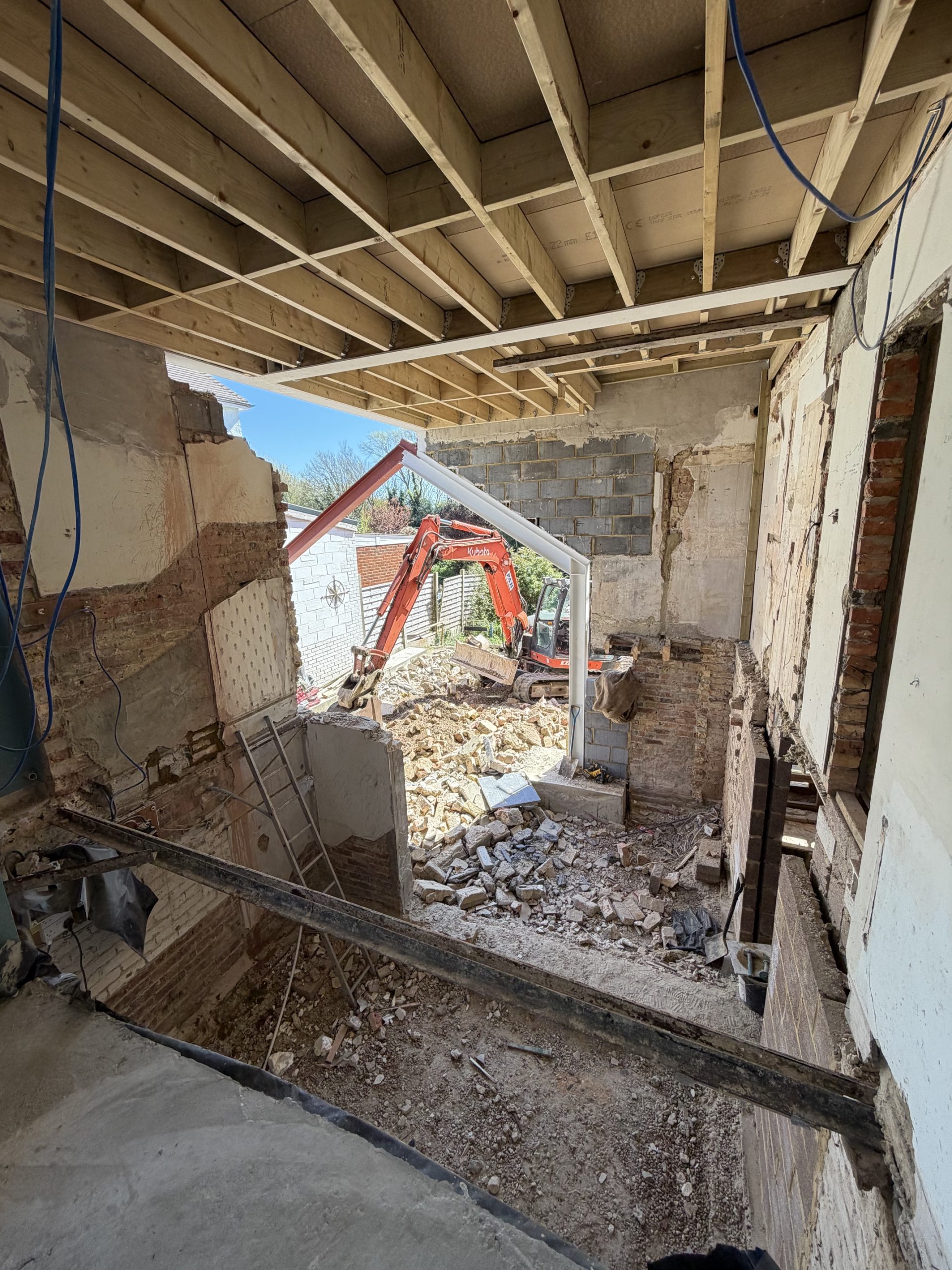 A partially demolished building with exposed brick walls and wooden beams, debris scattered on the ground, and a red excavator working outside under a metal framework.