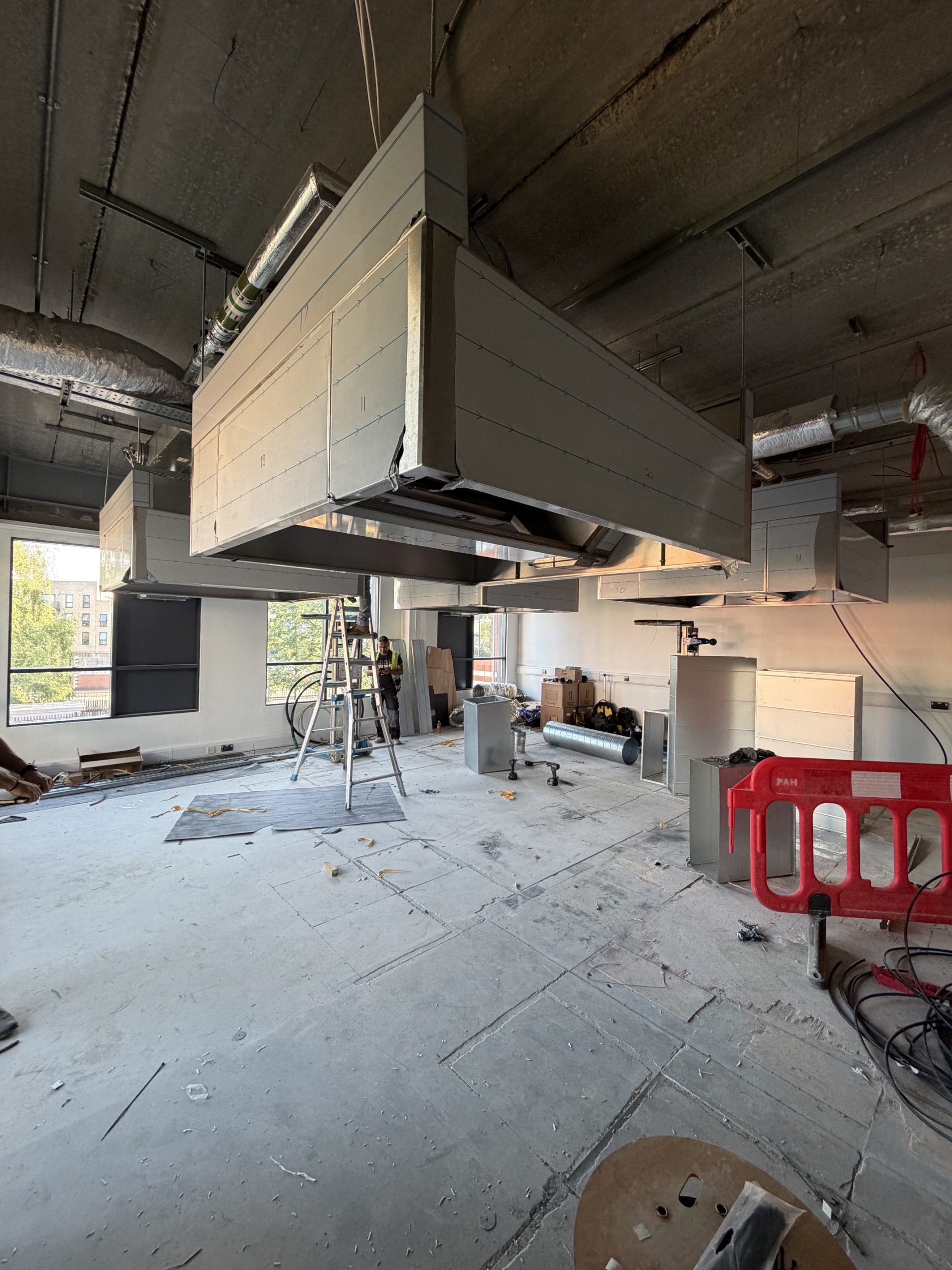 An unfinished room with exposed ceilings, large suspended ventilation units, construction materials, ladders, and open windows letting in daylight. The floor is covered with dust and debris from ongoing renovation work.