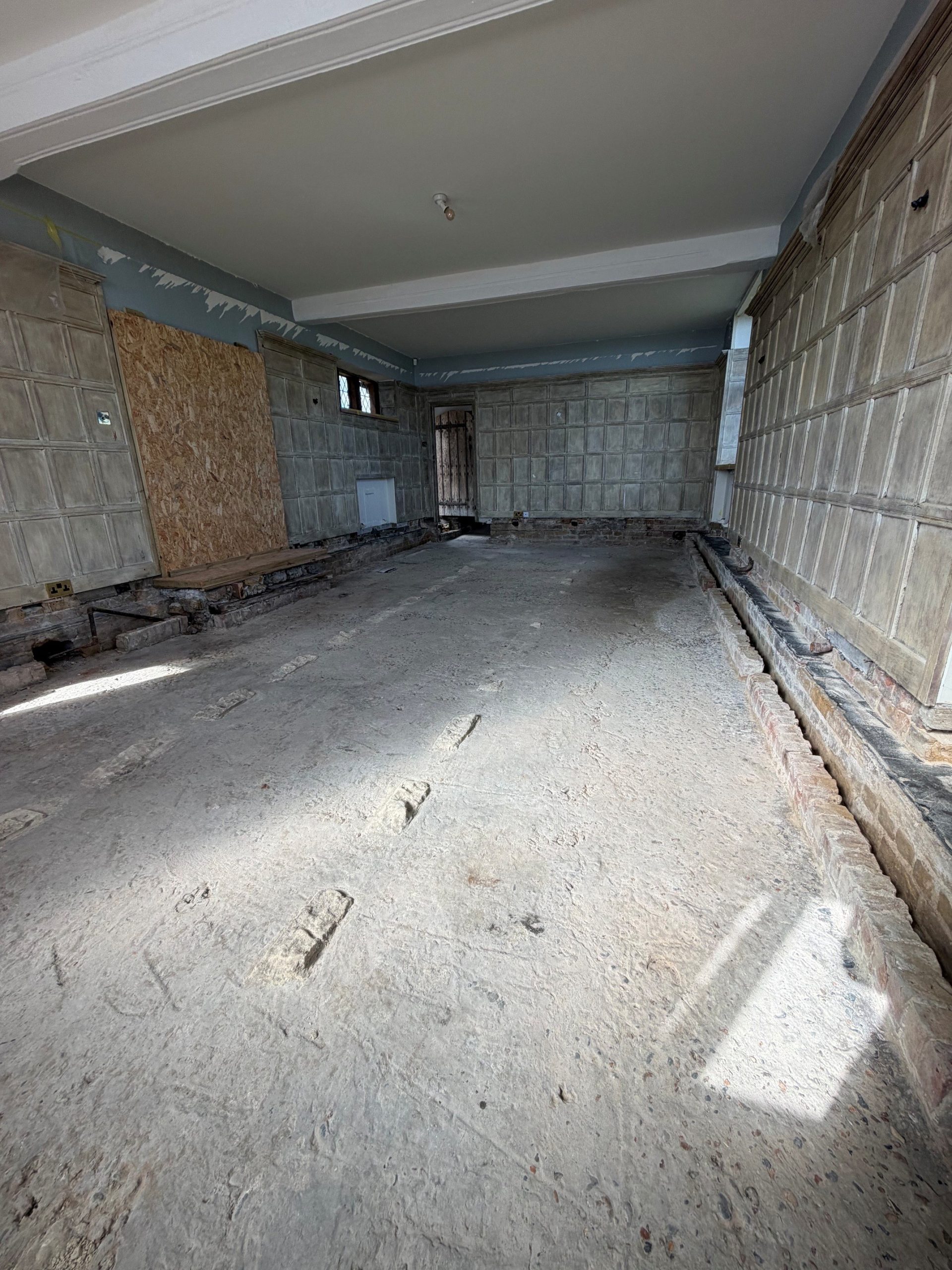 A large, empty room under renovation with exposed subfloor, unfinished walls, a boarded window, and visible construction materials along the sides. Light enters from a window on the right.