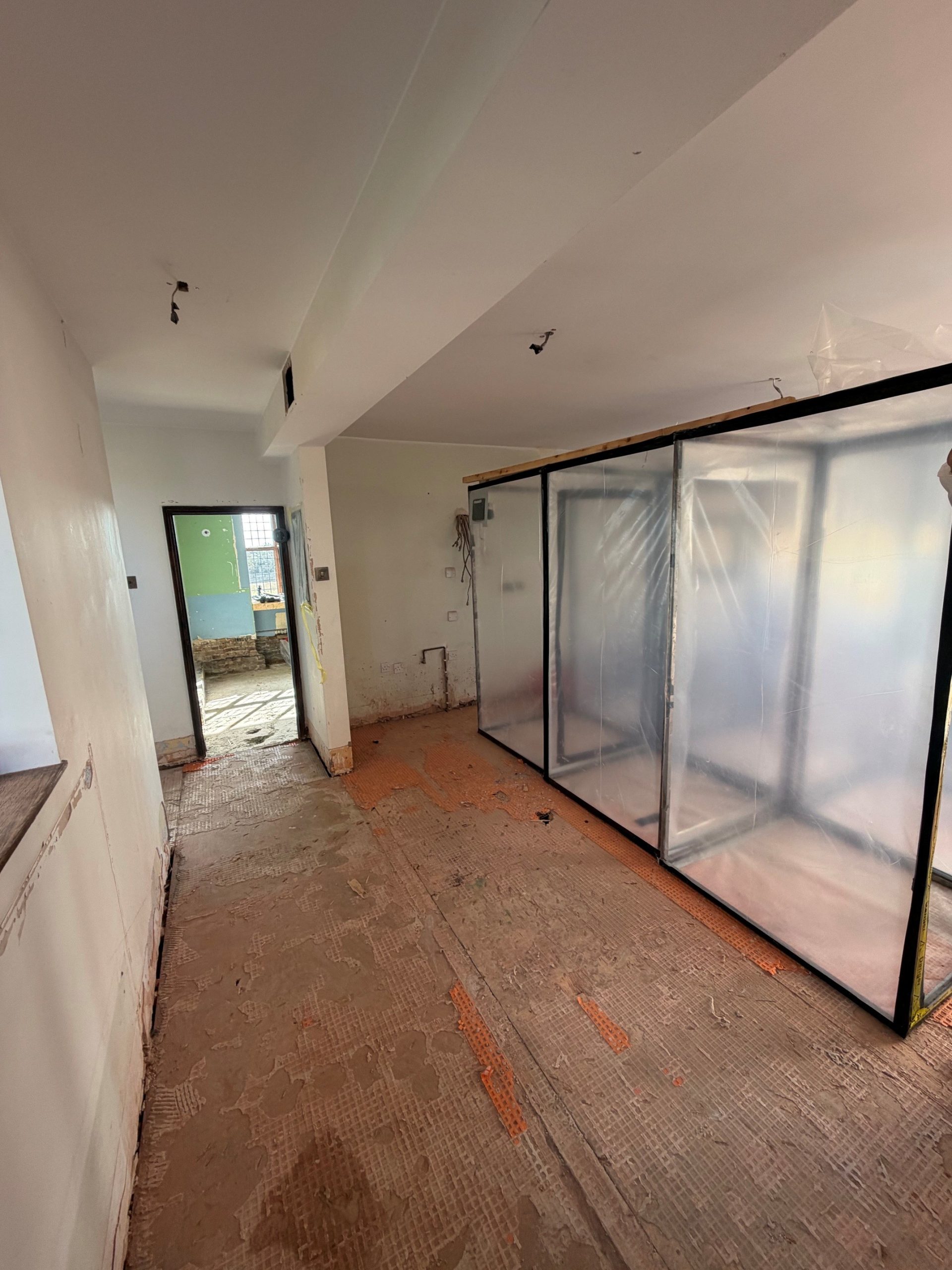 A hallway under renovation, with exposed subfloor and a large plastic sheet barrier on the right. Wires hang from the ceiling, and there is debris scattered on the floor. Light enters from a window and an open doorway in the background.