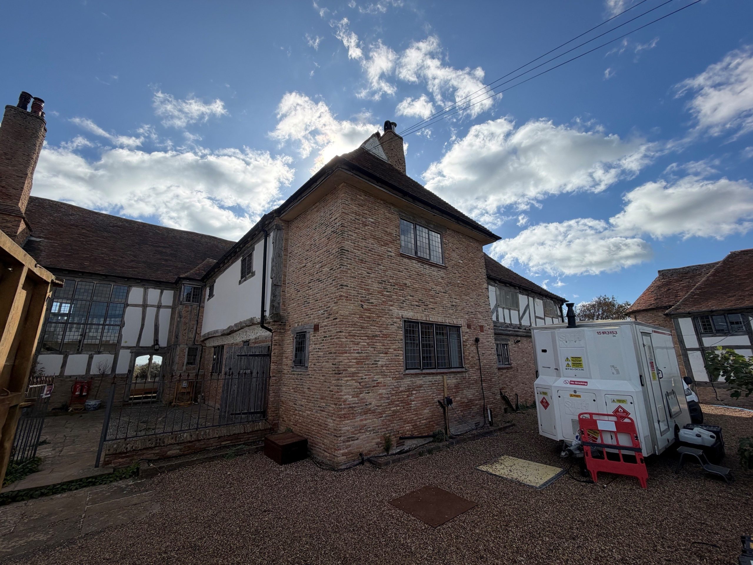 A brick and timber-framed building with a cloudy sky above, surrounded by gravel, a white utility unit, and red safety barriers in the yard.
