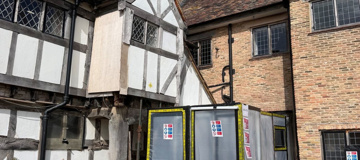 A historic half-timbered building with brick walls has a section cordoned off by plastic barriers and warning signs, likely due to construction or hazardous materials. The sky is clear and blue.