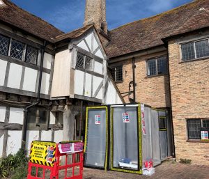 A historic half-timbered building with brick walls has a section cordoned off by plastic barriers and warning signs, likely due to construction or hazardous materials. The sky is clear and blue.