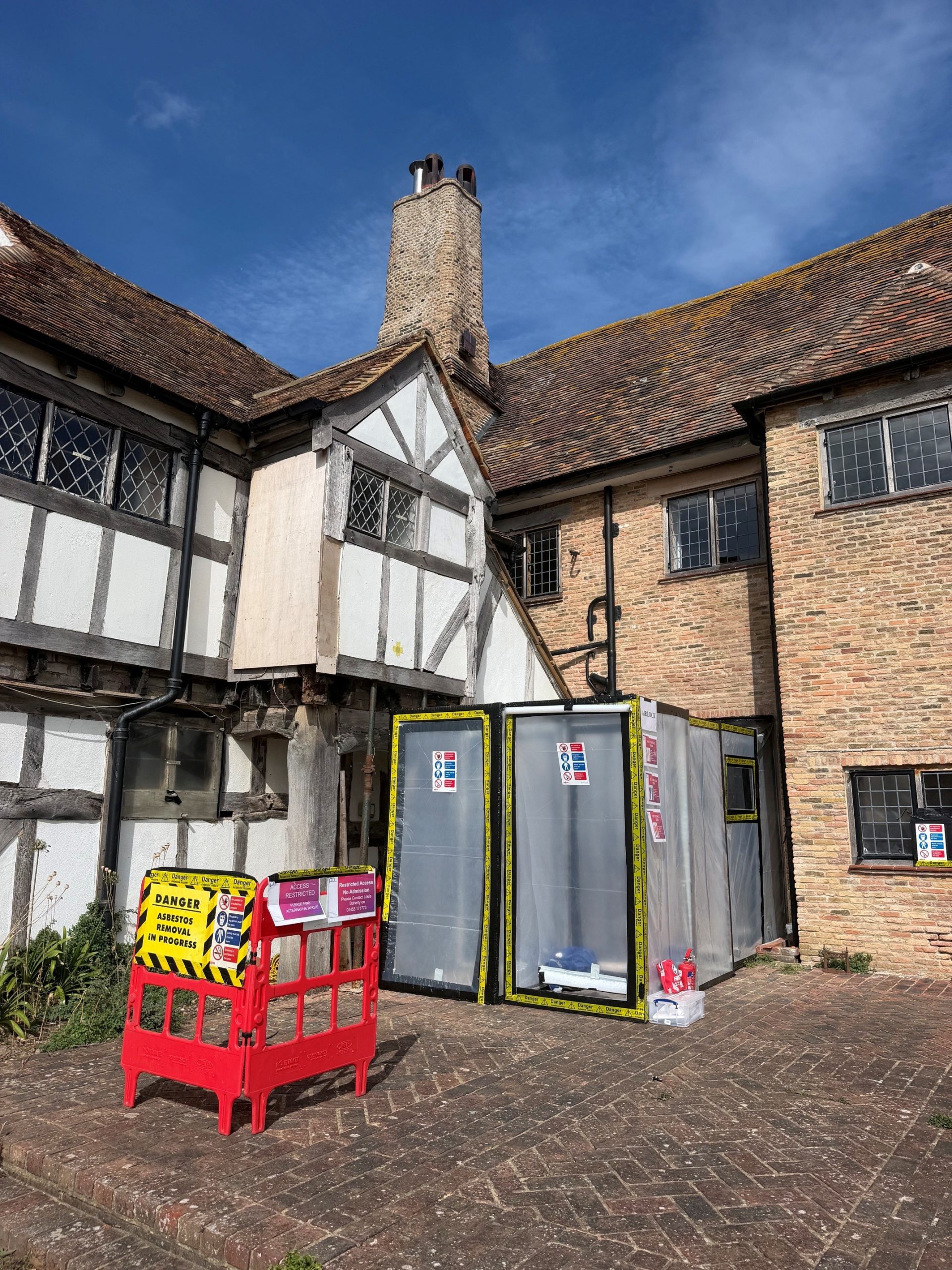 A historic half-timbered building with brick walls has a section cordoned off by plastic barriers and warning signs, likely due to construction or hazardous materials. The sky is clear and blue.