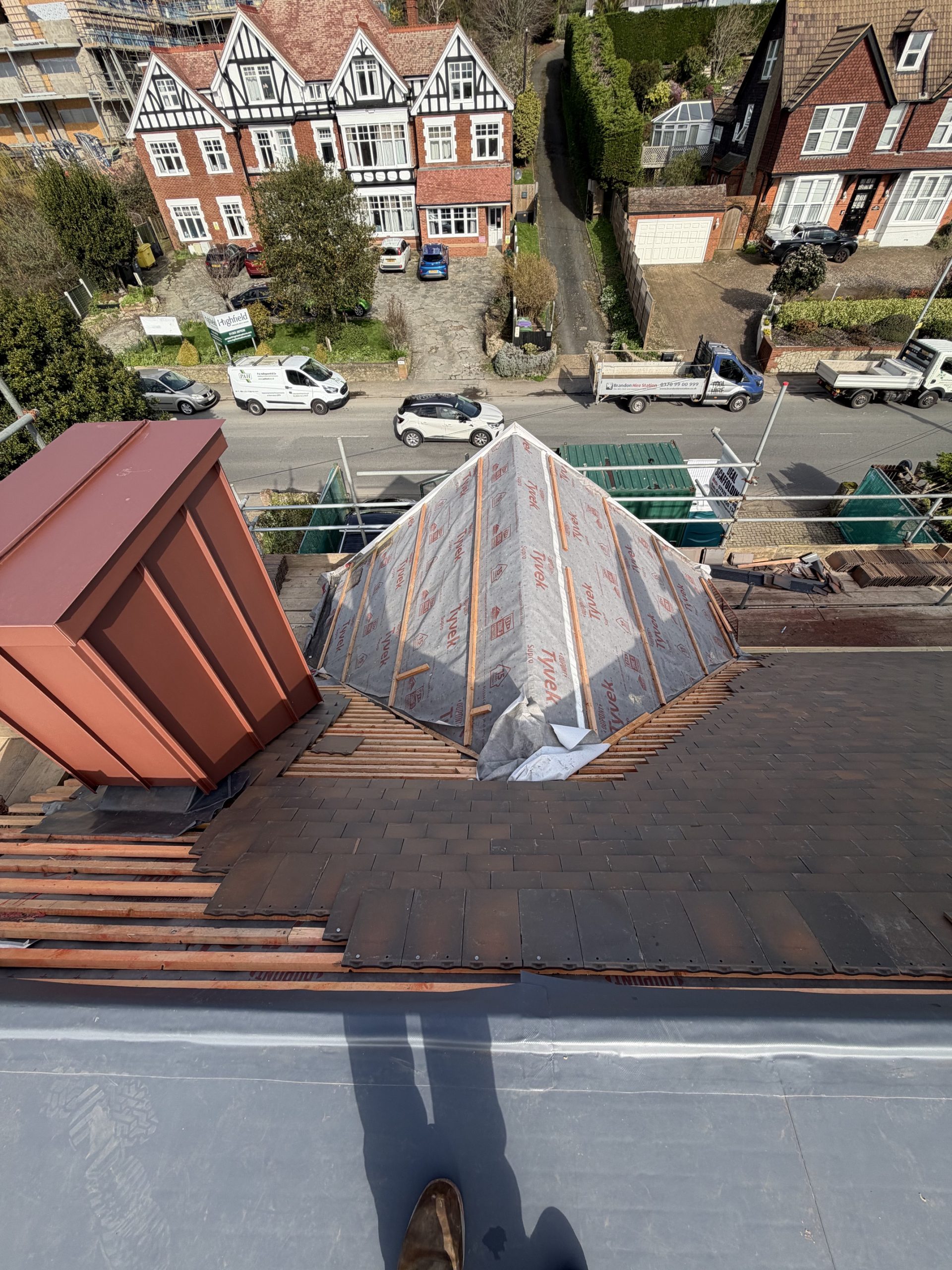 A rooftop under construction, partly covered with roofing felt and tiles. Scaffolding surrounds the roof. Residential houses, cars, and trees are visible on the street below. A shadow of a person appears in the foreground.