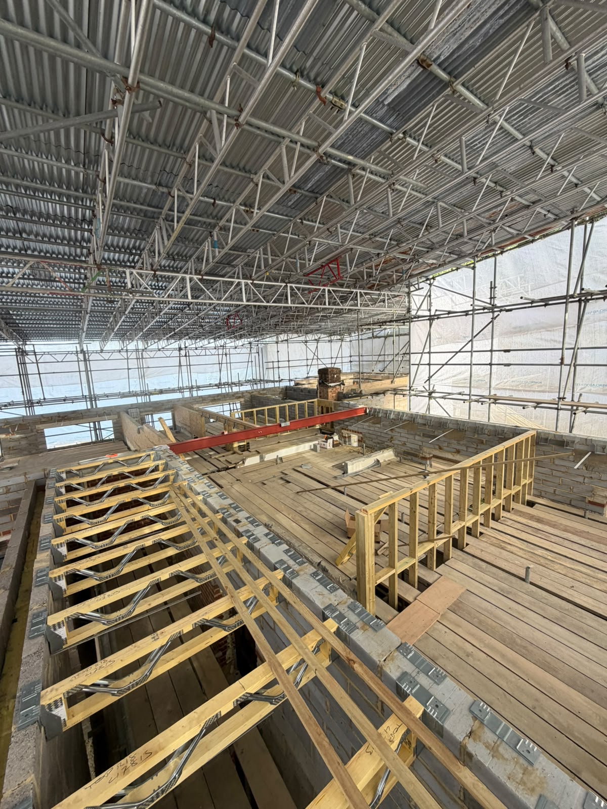 Indoor construction site with extensive scaffolding overhead, wooden beams and planks on the floor, partially built walls, and stacks of building materials throughout the area.