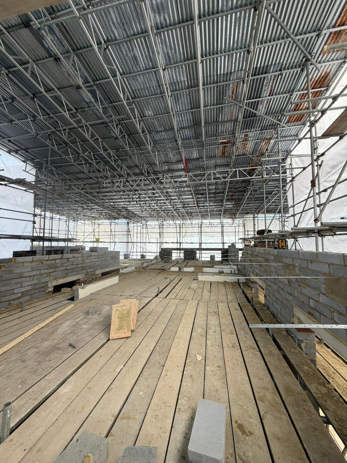 Construction site with metal scaffolding overhead, wooden planks on the floor, and stacks of concrete blocks arranged throughout the partially built structure. Bright lighting suggests work is happening during the day.